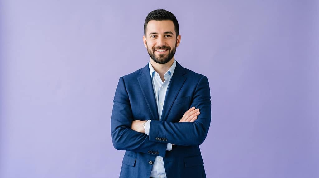 : Confident smiling young businessman in navy suit standing with crossed arms against purple background